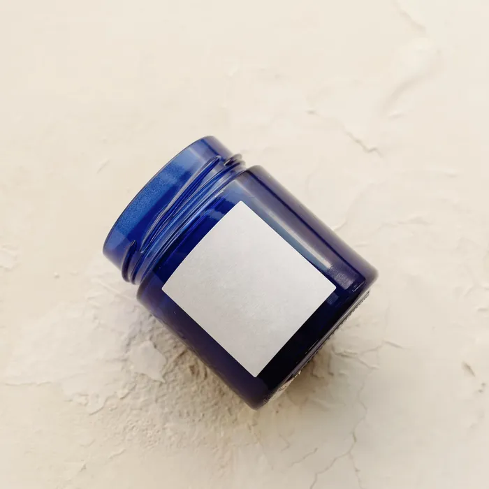 A small blue glass jar lying on a plaster surface with a blank white label on it