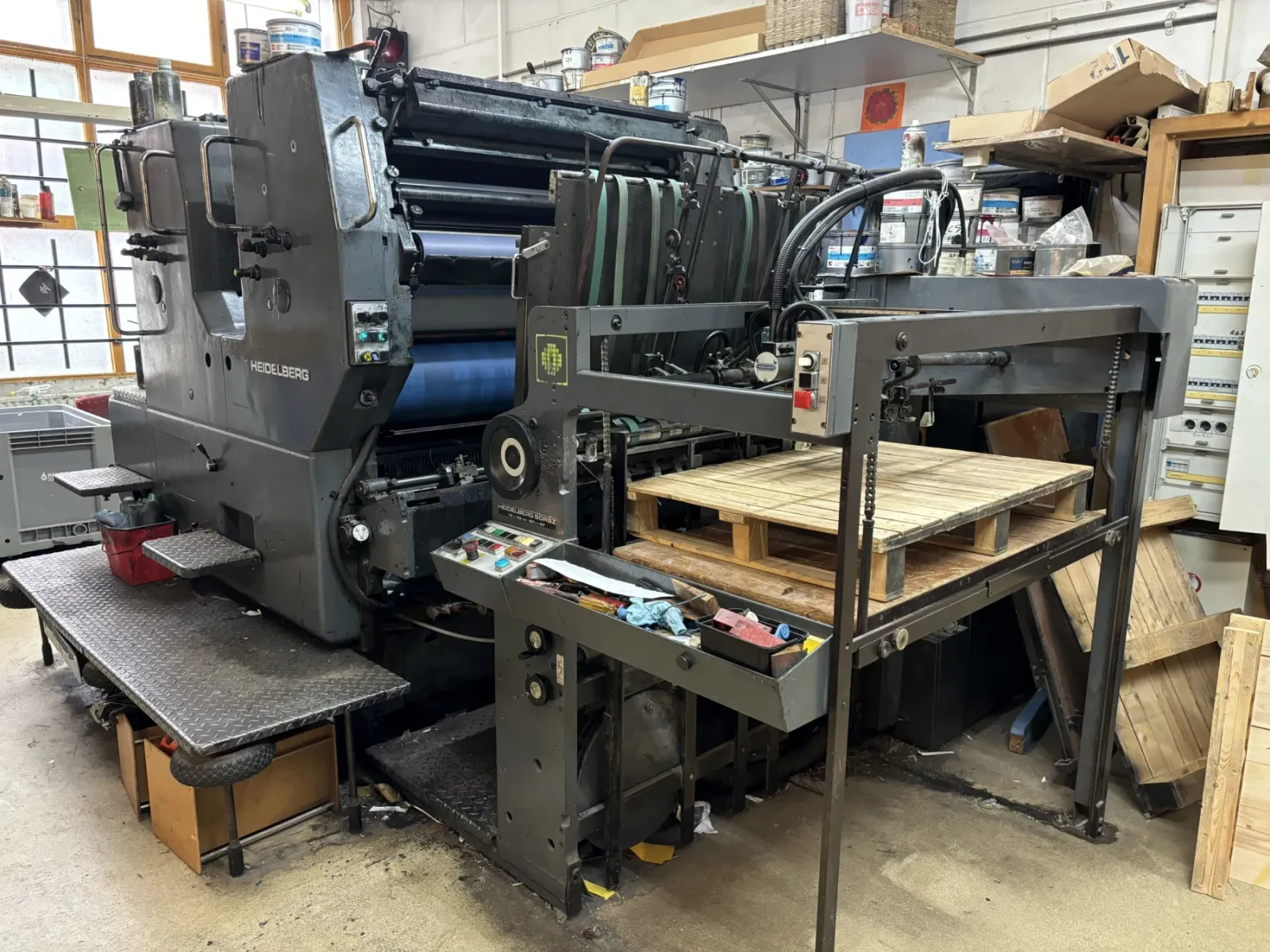 A sheet-fed offset press with ink tins stacked on shelving behind it